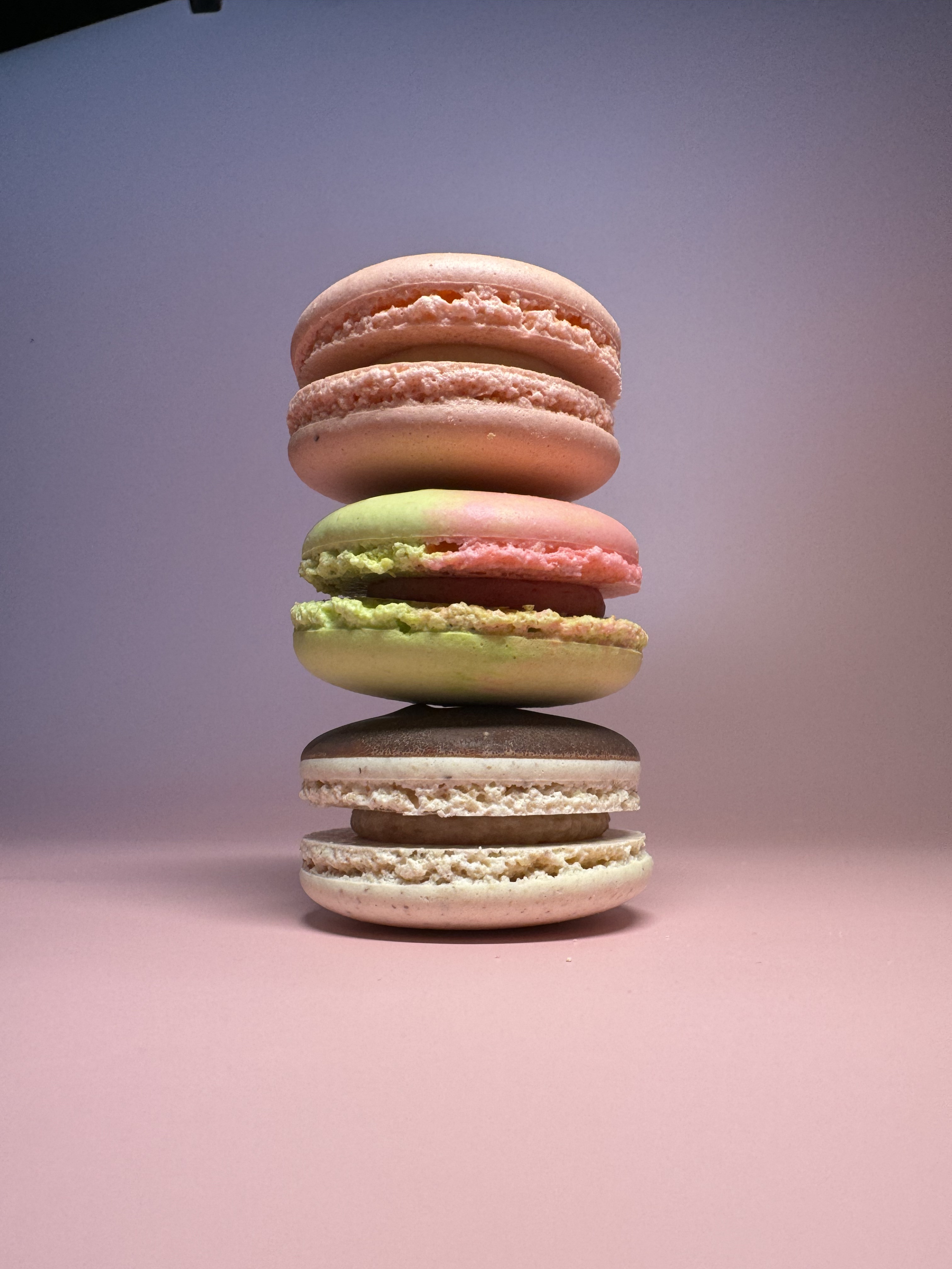 three macaron cookies are stacked on top of eachother, each is a different color, one is pink, one is green and pink, one is blonde and brown, the background of the image is light purple